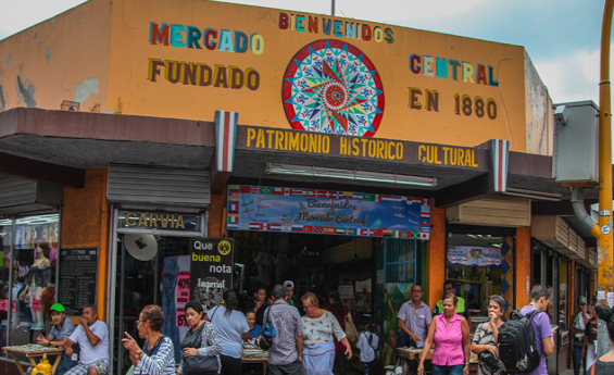 Se muestra imagen de la entrada principal del Mercado Central del Cantón de San José. En la parte superior de la imagen cuyo fondo es en tonos amarillo con leyenda que dice bienvenidos Mercado Central fundado en 1880. Además contiene imágen de una carreta típica costarricense y leyenda Patrimonio Histórico Cultural. Hay grupo de personas transeúntes que ingresan, salen o pasan al frente de la entrada principal. 